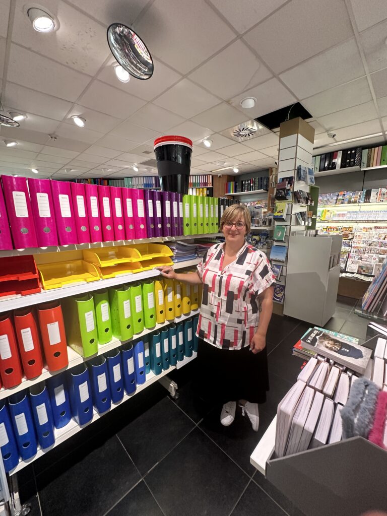 Isabelle, propriétaire de la librairie-presse Paperas à Gerpinnes, debout près d'une étagère colorée de classeurs et fournitures de bureau.