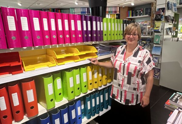 Isabelle, propriétaire de la librairie-presse Paperas à Gerpinnes, debout près d'une étagère colorée de classeurs et fournitures de bureau.